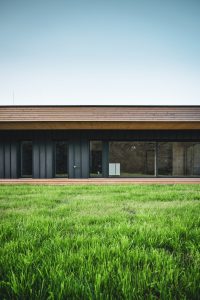 Detail přízemního domu s tmavou fasádou, velkým oknem a technickou jednotkou u stěny