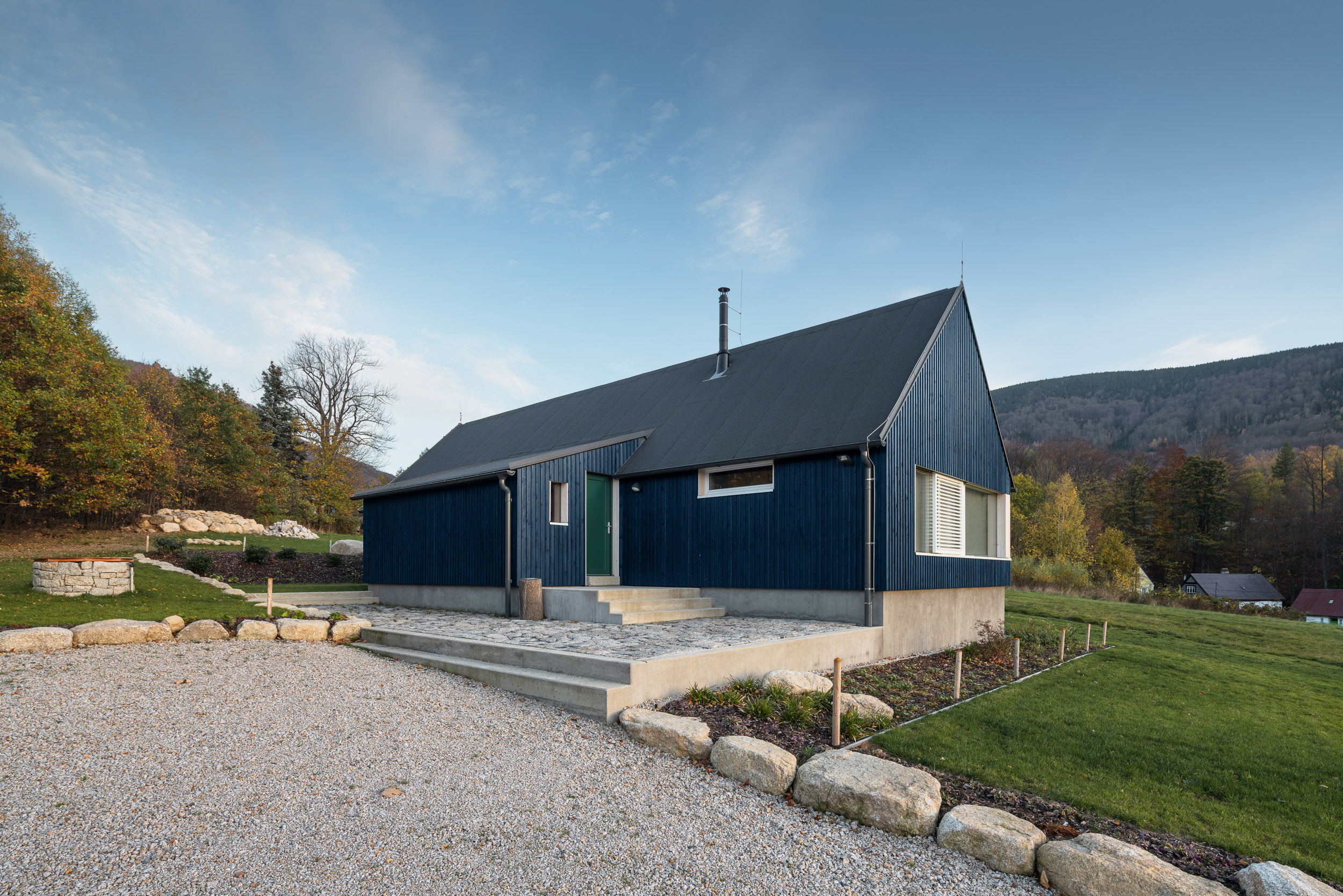 Vstupní část chaty s modrou fasádou, betonovým soklem a kamenným lemováním postavena firmou 3AE