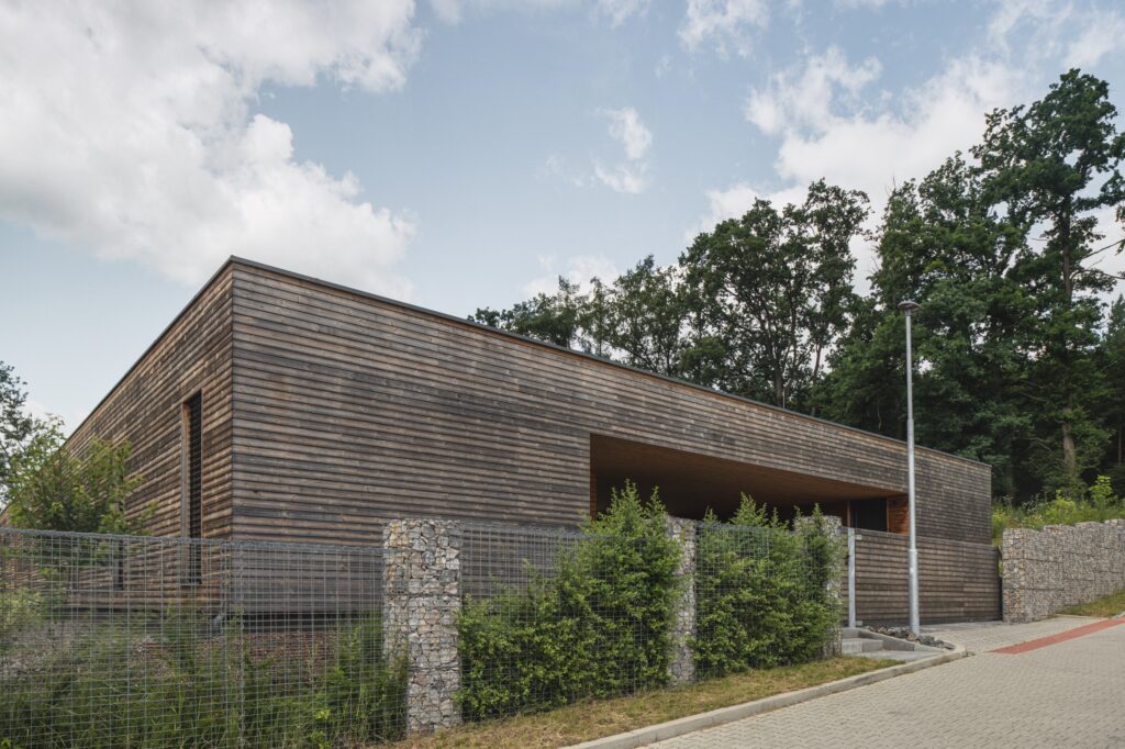 Pohled na vstupní část dřevostavby s dřevěnou fasádou a gabionovým plotem